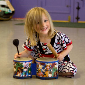 A grinning blond little girl in a bright outfit bangs on colorful bongos