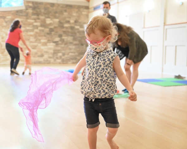 A child in red glasses and a bright shirt dances with a pink scarf, while other children and parents dance in the background